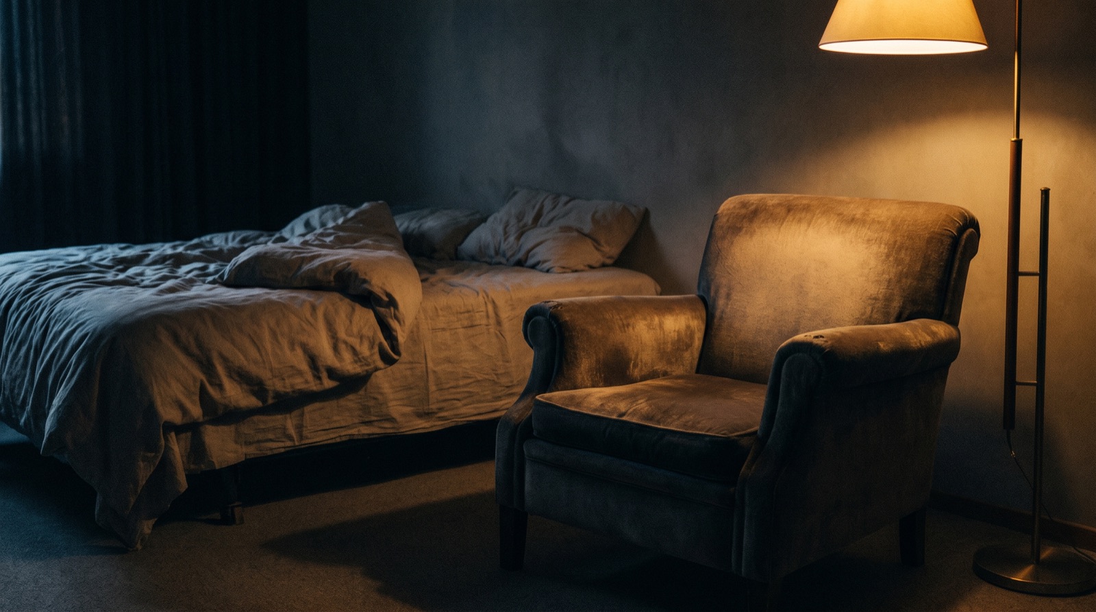 Ein ungemachtes Bett und ein abgenutzter Sessel in einem dämmrigen Zimmer, daneben eine brennende Stehlampe